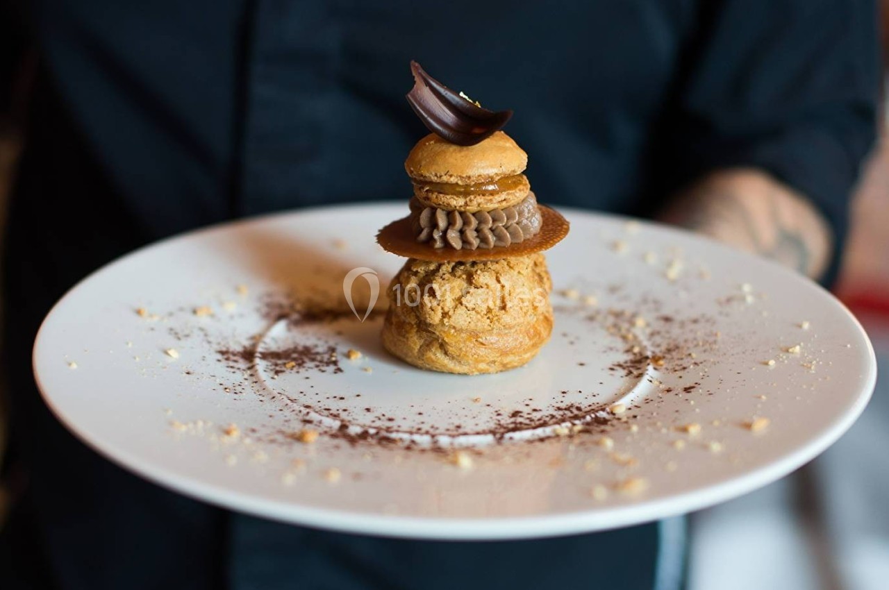 Pâtisserie élégante servie sur une assiette blanche, décorée de cacao en poudre et tenue par une personne en arrière-plan.