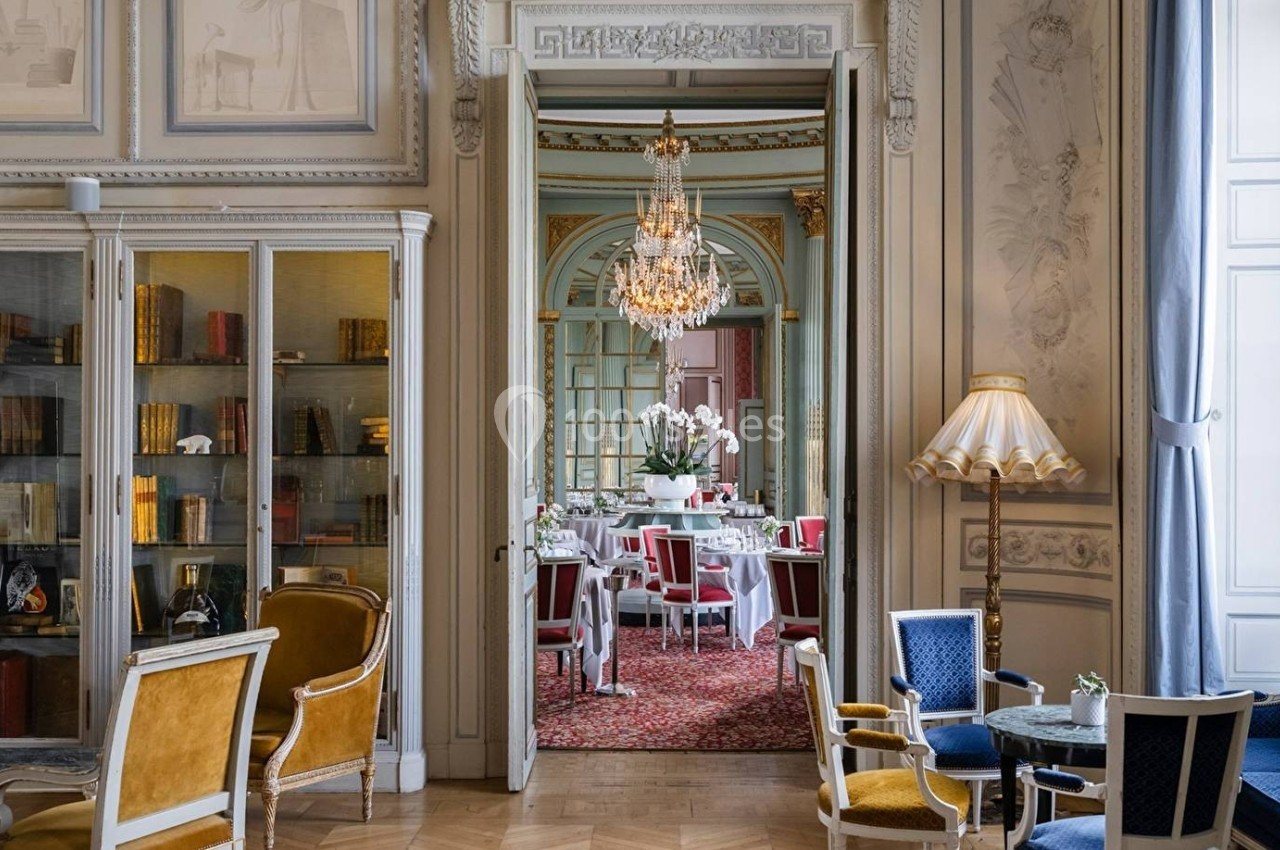 Salon élégant avec mobilier classique, bibliothèque vitrée, et vue sur une salle à manger ornée d'un lustre.