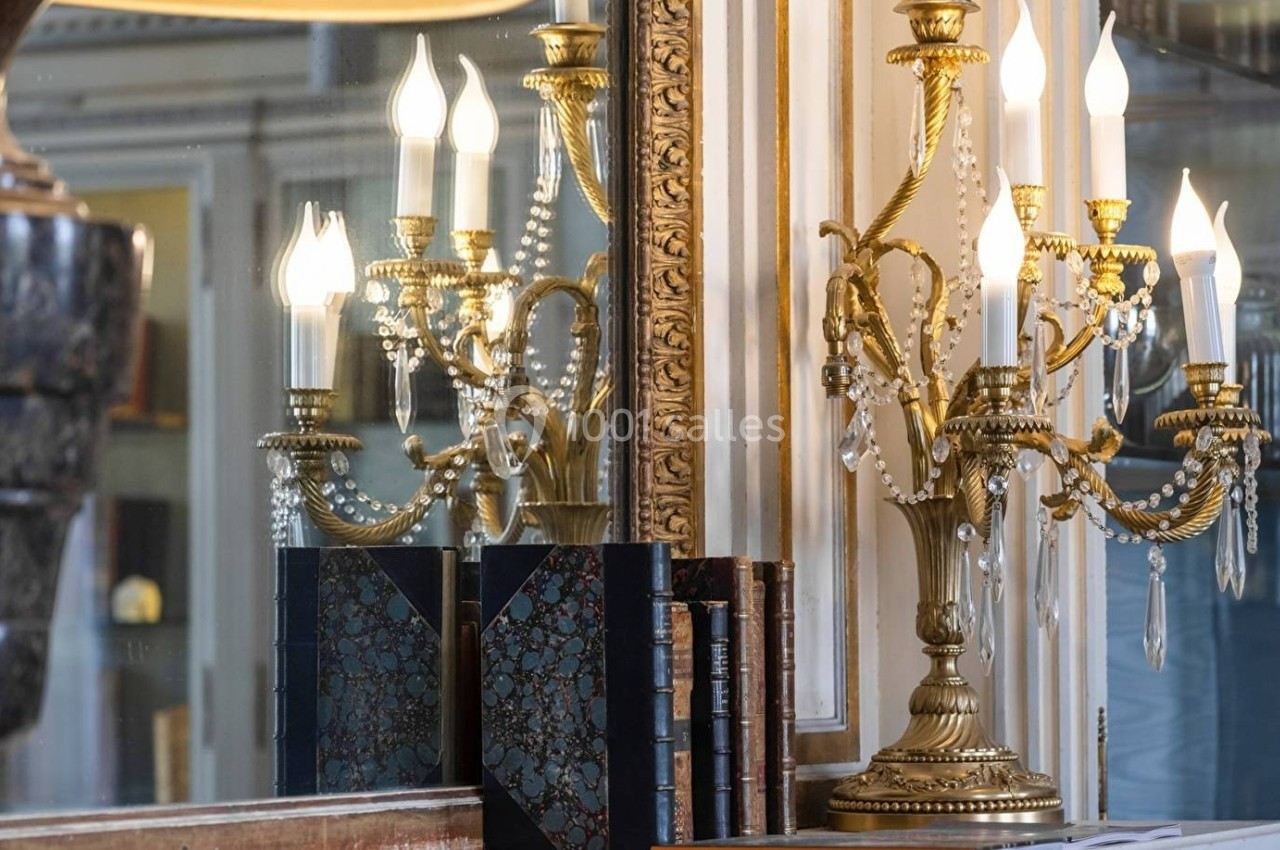 Chandelier doré à plusieurs branches avec des ampoules allumées, reflété dans un miroir, posé près de livres anciens.