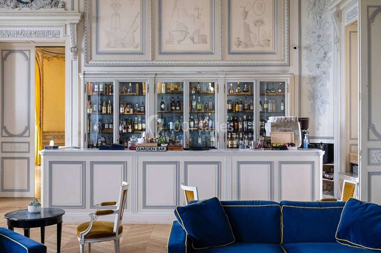 Bar élégant avec comptoir blanc, étagères remplies de bouteilles, fauteuils bleus et décoration intérieure classique.