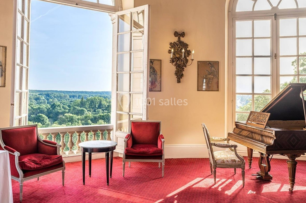 Salon lumineux avec deux fauteuils rouges, une table, un piano à queue et une vue sur un paysage verdoyant à travers de…
