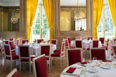 Salle à manger élégante avec table dressée, chaises rouges, murs dorés, lustres et grandes fenêtres avec rideaux jaunes.