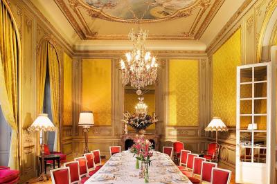 Salle à manger élégante avec table dressée, chaises rouges, murs dorés, lustres et grandes fenêtres avec rideaux jaunes.