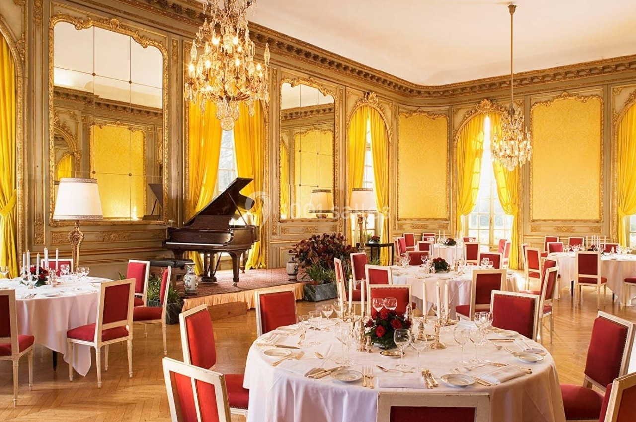 Salle élégante avec tables dressées, chaises rouges, rideaux jaunes, miroirs dorés et un piano à queue sur une estrade.