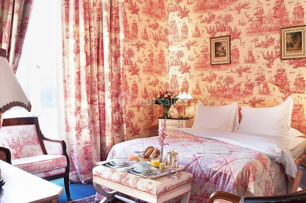 Chambre décorée de motifs toile de Jouy rouges avec lit double, table de petit-déjeuner et lumière naturelle.