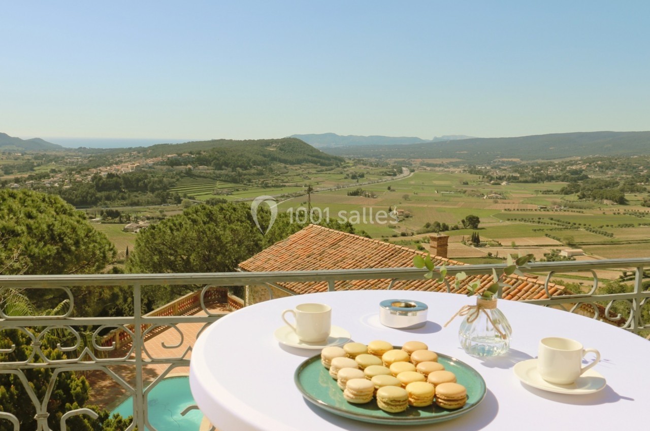 Table avec macarons, tasses de café et vase, offrant une vue panoramique sur une vallée ensoleillée et verdoyante.
