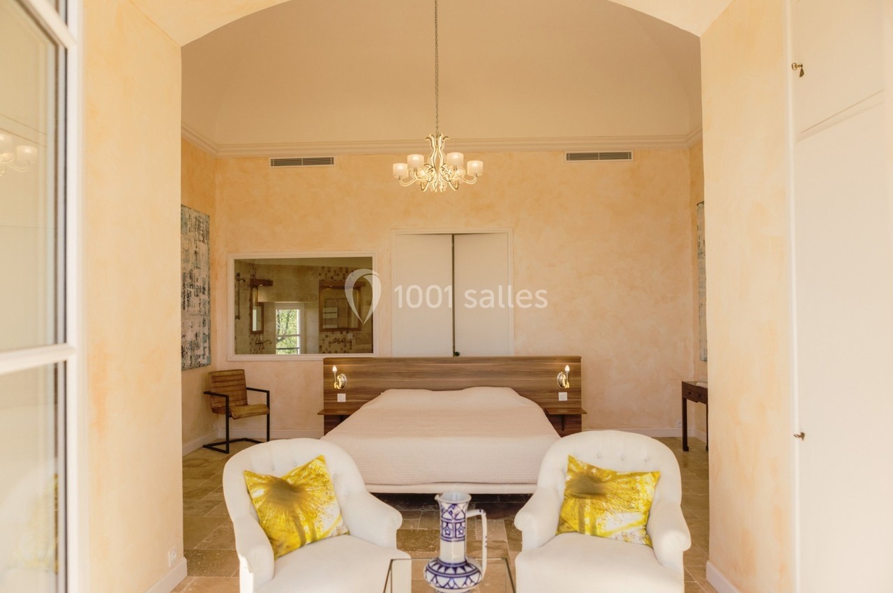 Chambre lumineuse avec un lit double, deux fauteuils blancs, coussins jaunes et un lustre suspendu au plafond.