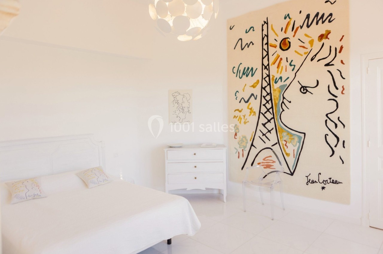 Chambre lumineuse avec un lit double, une commode blanche et une grande œuvre murale colorée représentant une tour et un…