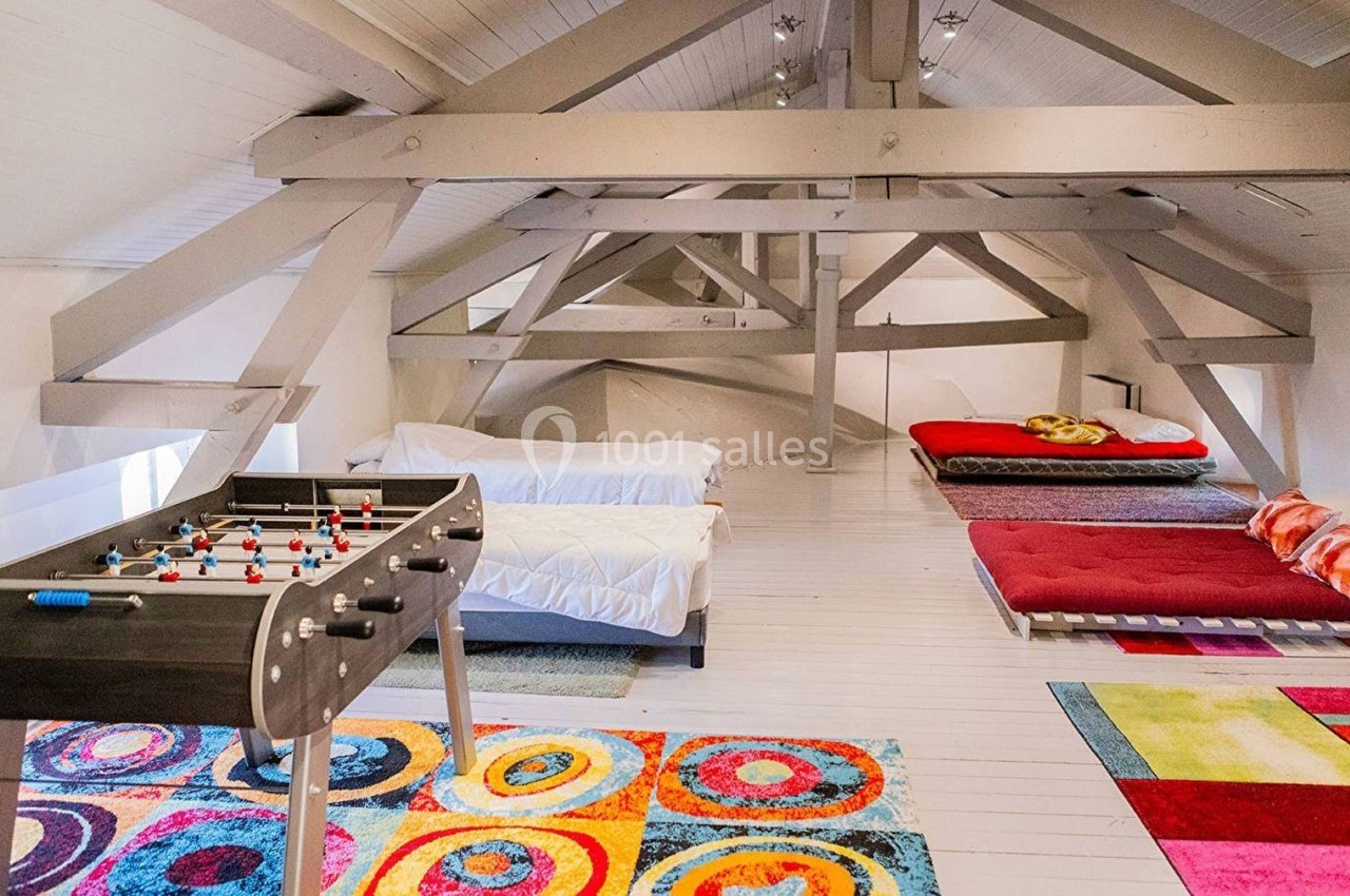 Grenier aménagé avec lits, baby-foot et tapis colorés, sous une charpente apparente en bois peint.