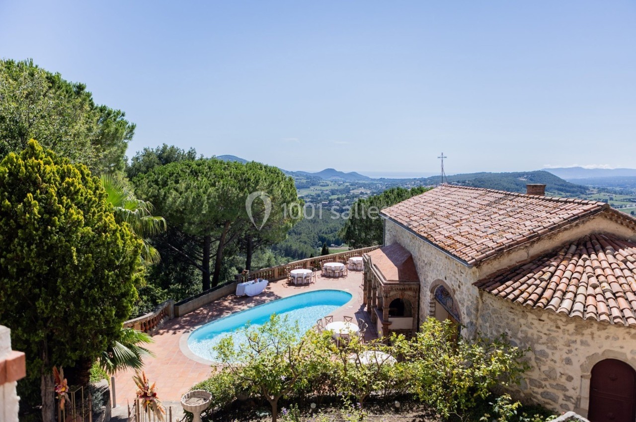 Vue d'une maison en pierre avec toit en tuiles, piscine et terrasse, entourée de végétation et paysage vallonné.