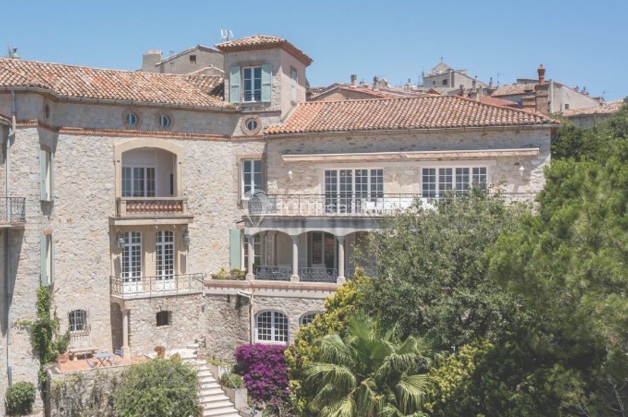 Façade d'une grande maison en pierre avec balcons, entourée de végétation et baignée de lumière sous un ciel bleu.