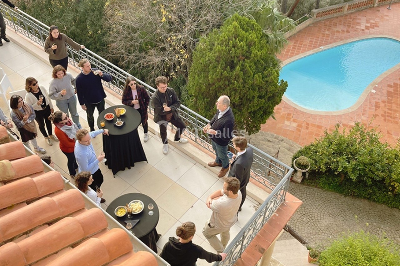 Groupe de personnes discutant autour de tables hautes sur une terrasse surplombant une piscine.