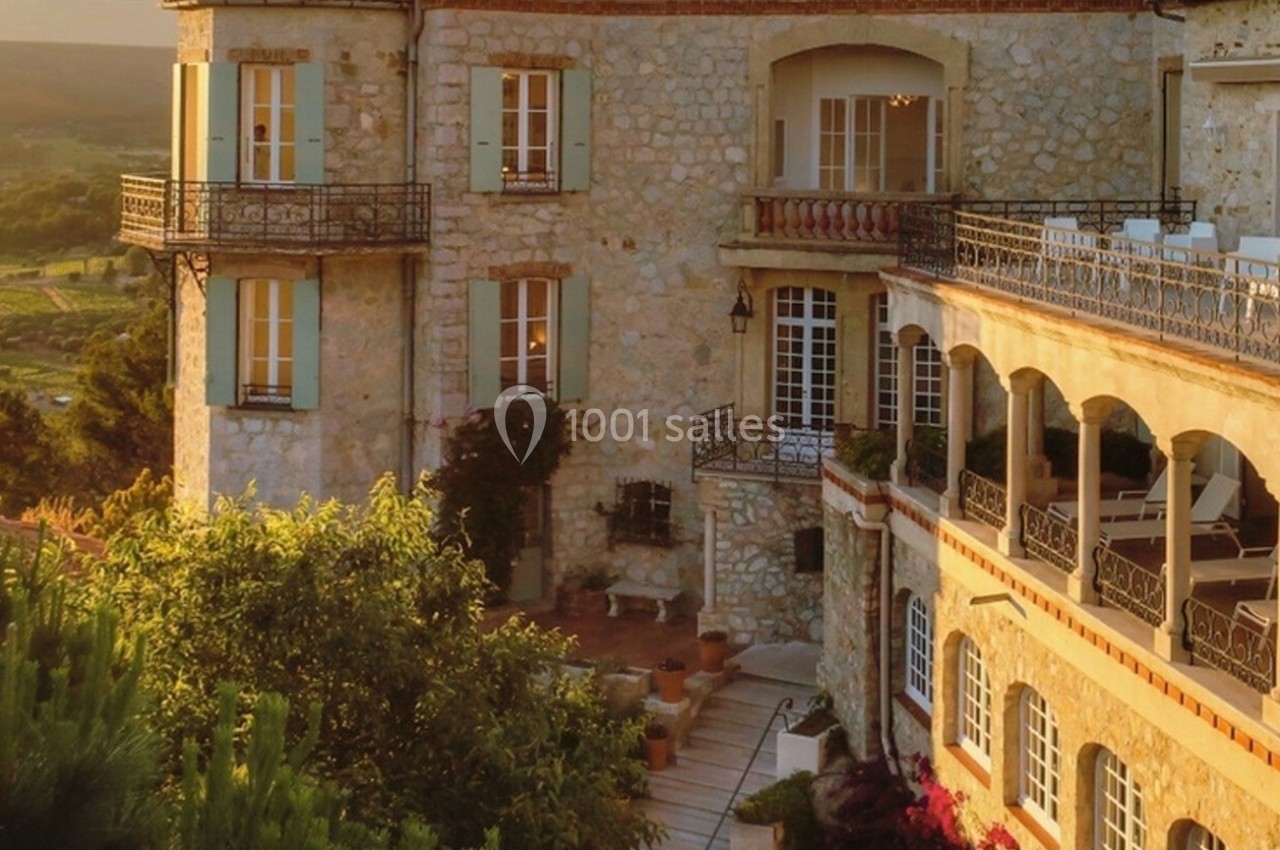 Façade en pierre d'un bâtiment avec balcons et terrasses, entouré de végétation sous une lumière dorée.