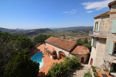 Vue d'une maison en pierre avec piscine, entourée de végétation, donnant sur une vallée et des collines sous un ciel bleu.