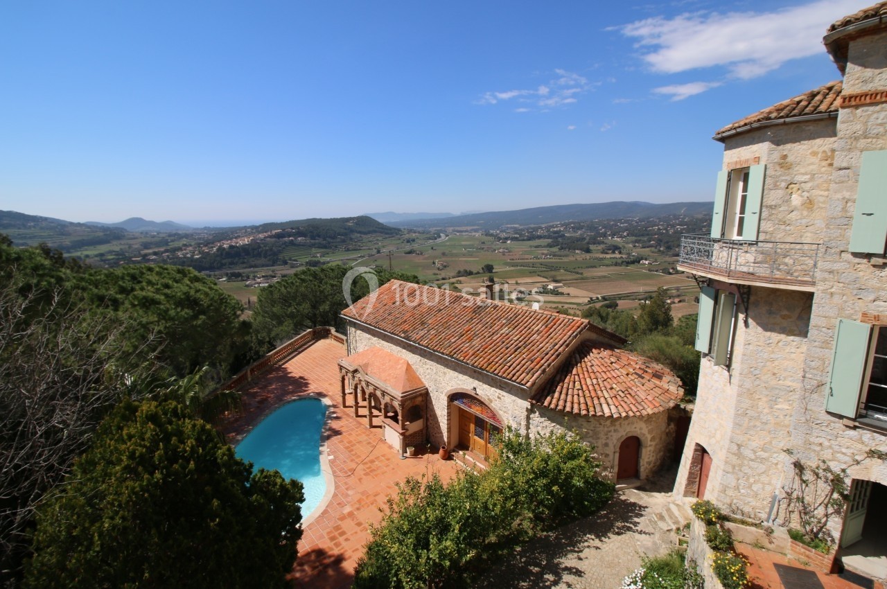Vue d'une maison en pierre avec piscine, entourée de végétation, donnant sur une vallée et des collines sous un ciel bleu.