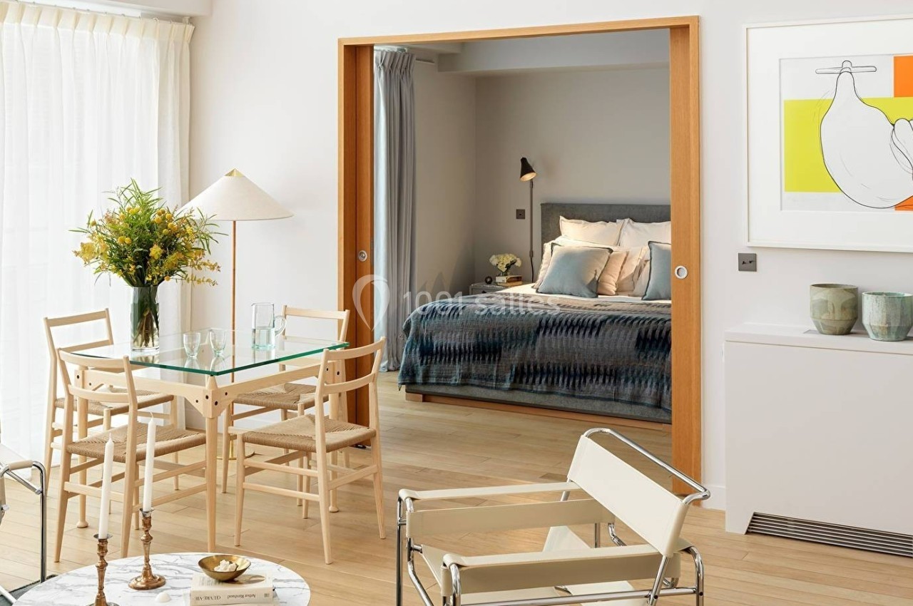 Salle à manger lumineuse avec table en verre, chaises en bois clair, et vue sur une chambre avec lit double.