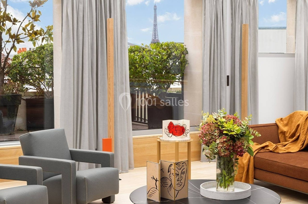 Salon lumineux avec fauteuils gris, canapé marron, table basse décorée et vue sur la Tour Eiffel à travers les fenêtres.