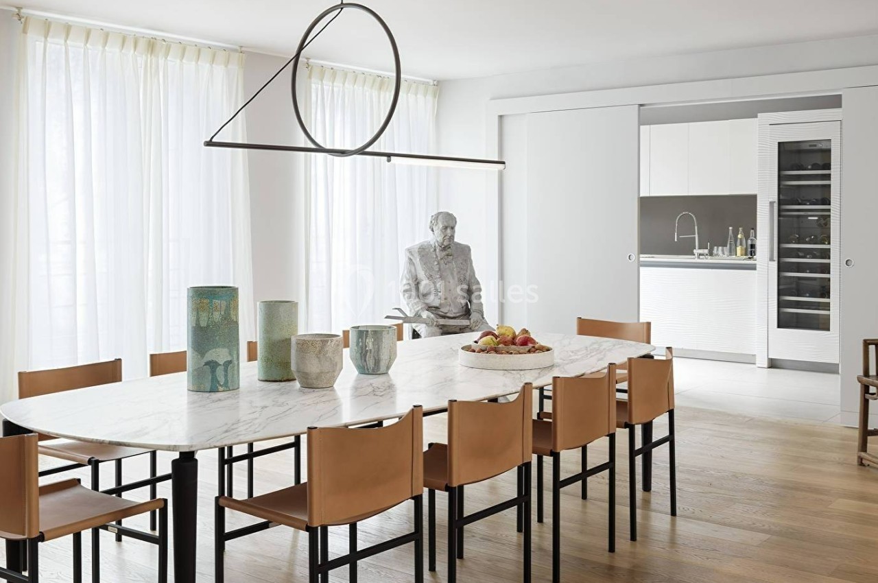 Salle à manger moderne avec table ovale en marbre, chaises en cuir, suspension design et cuisine ouverte en arrière-plan.