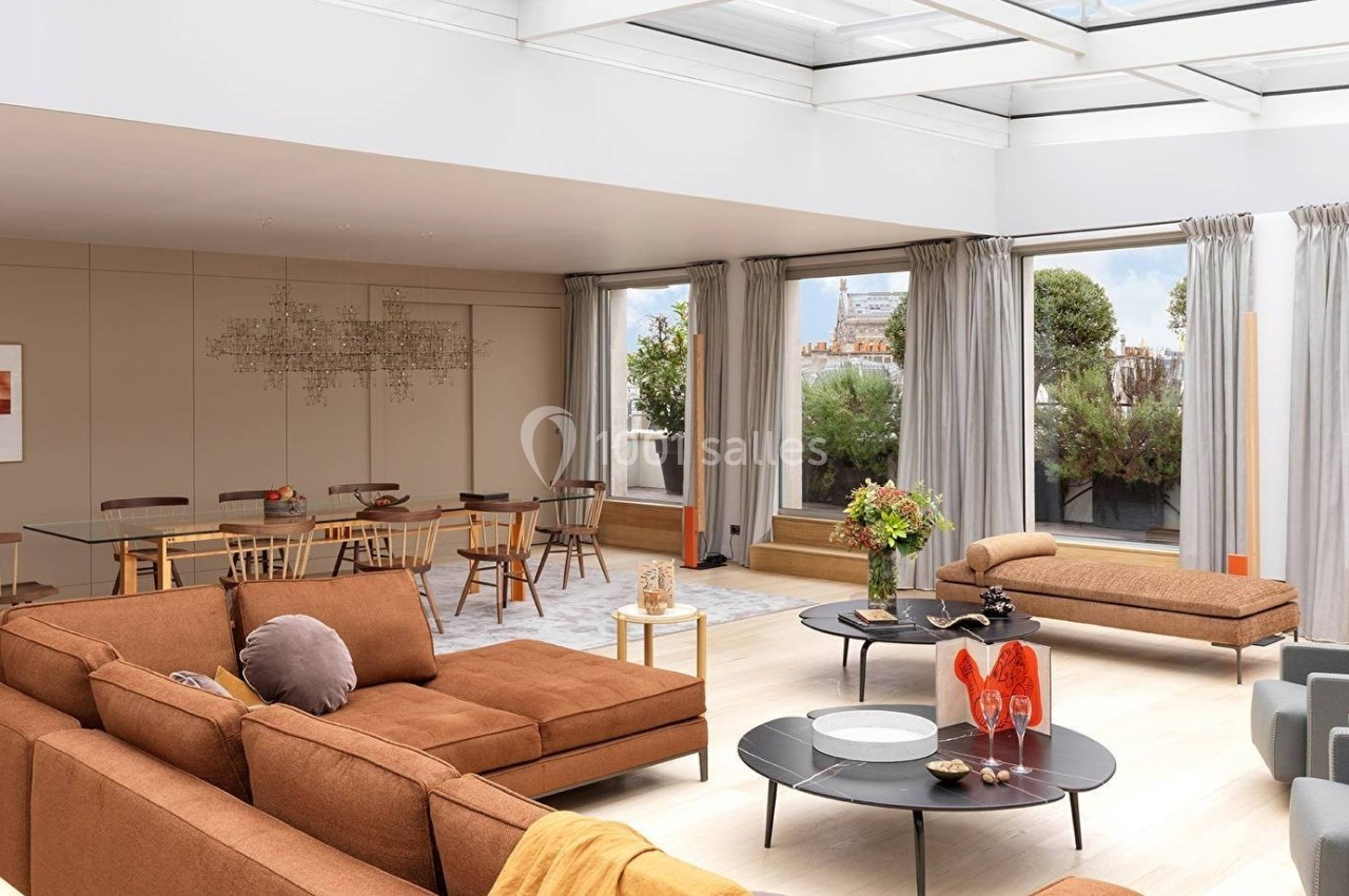 Salon lumineux avec canapé modulable brun, table à manger en verre, grandes baies vitrées et terrasse arborée.