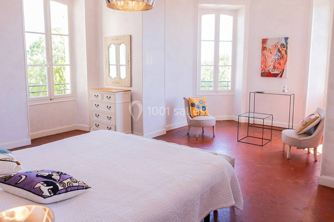 Chambre lumineuse avec lit blanc, mobilier clair, deux fenêtres, et décoration colorée sur les murs et les coussins.