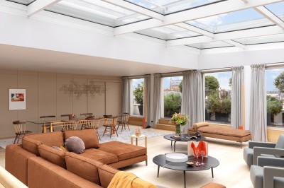 Salon lumineux avec canapé beige, table basse en bois, salle à manger attenante et escalier en arrière-plan.