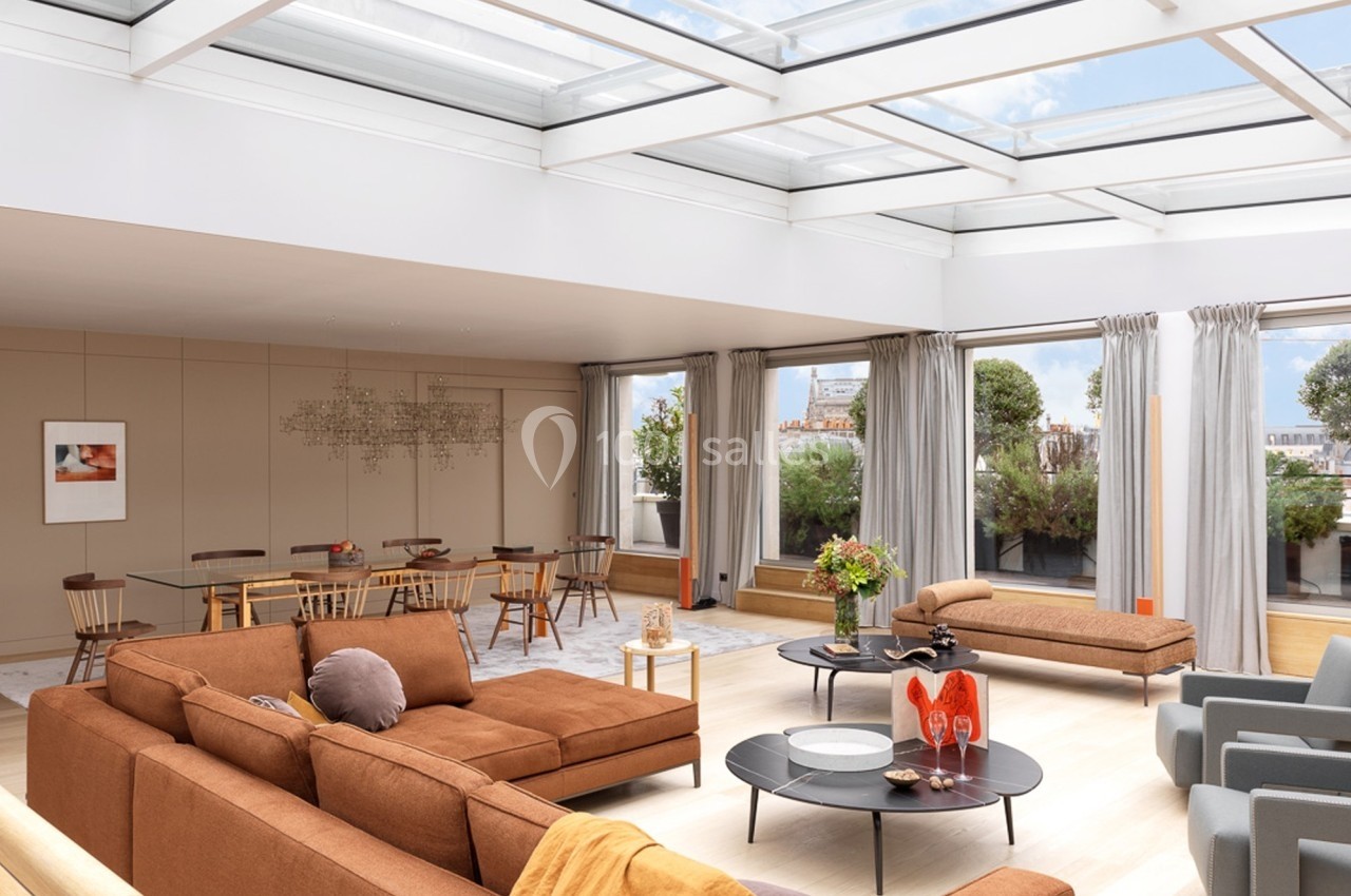 Salon lumineux avec toit vitré, mobilier moderne aux tons chauds, table à manger en arrière-plan et vue sur un jardin.