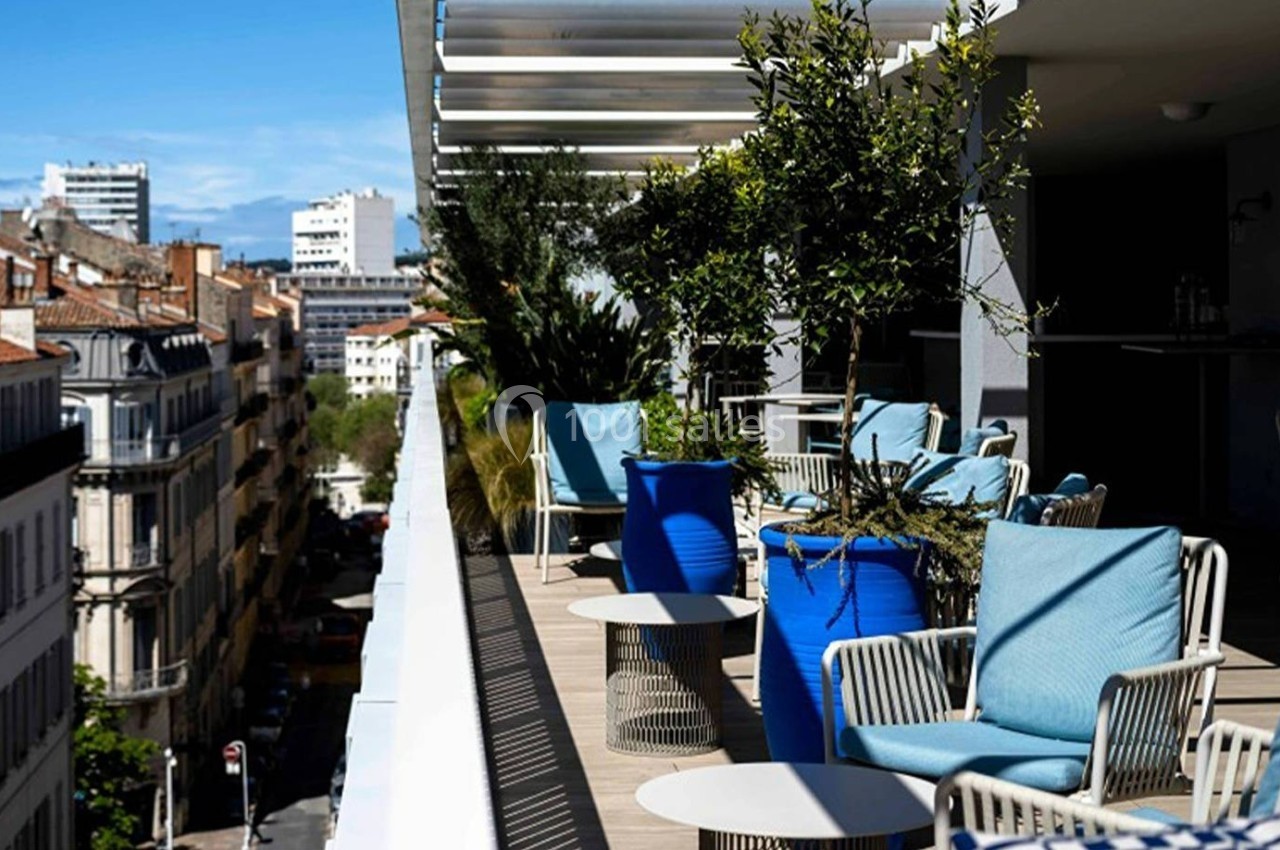 Terrasse ensoleillée avec fauteuils bleus, plantes en pots et vue sur des immeubles urbains sous un ciel dégagé.