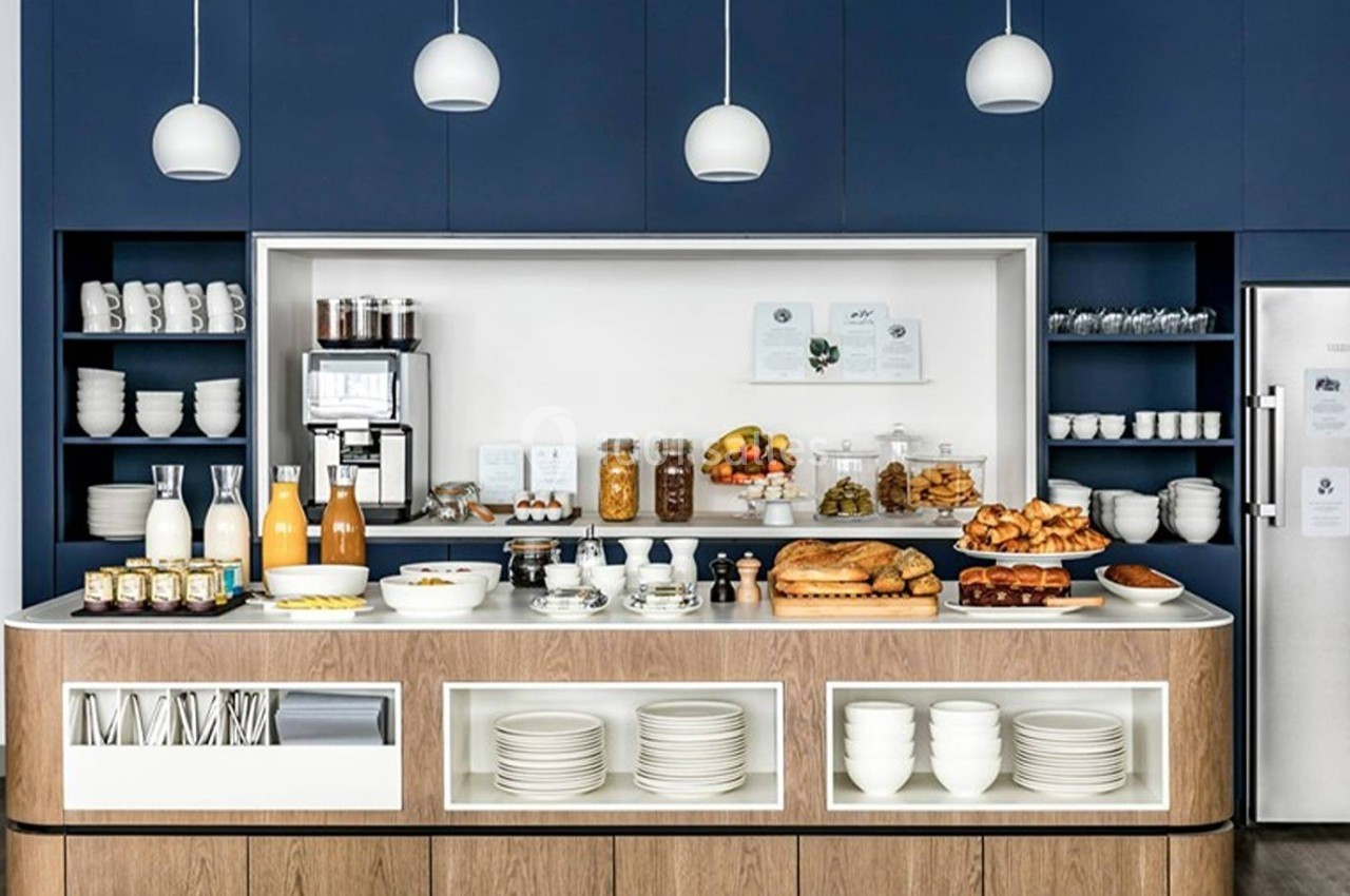 Buffet de petit-déjeuner avec boissons, viennoiseries, fruits et vaisselle, dans une cuisine moderne aux tons bleus et bois.