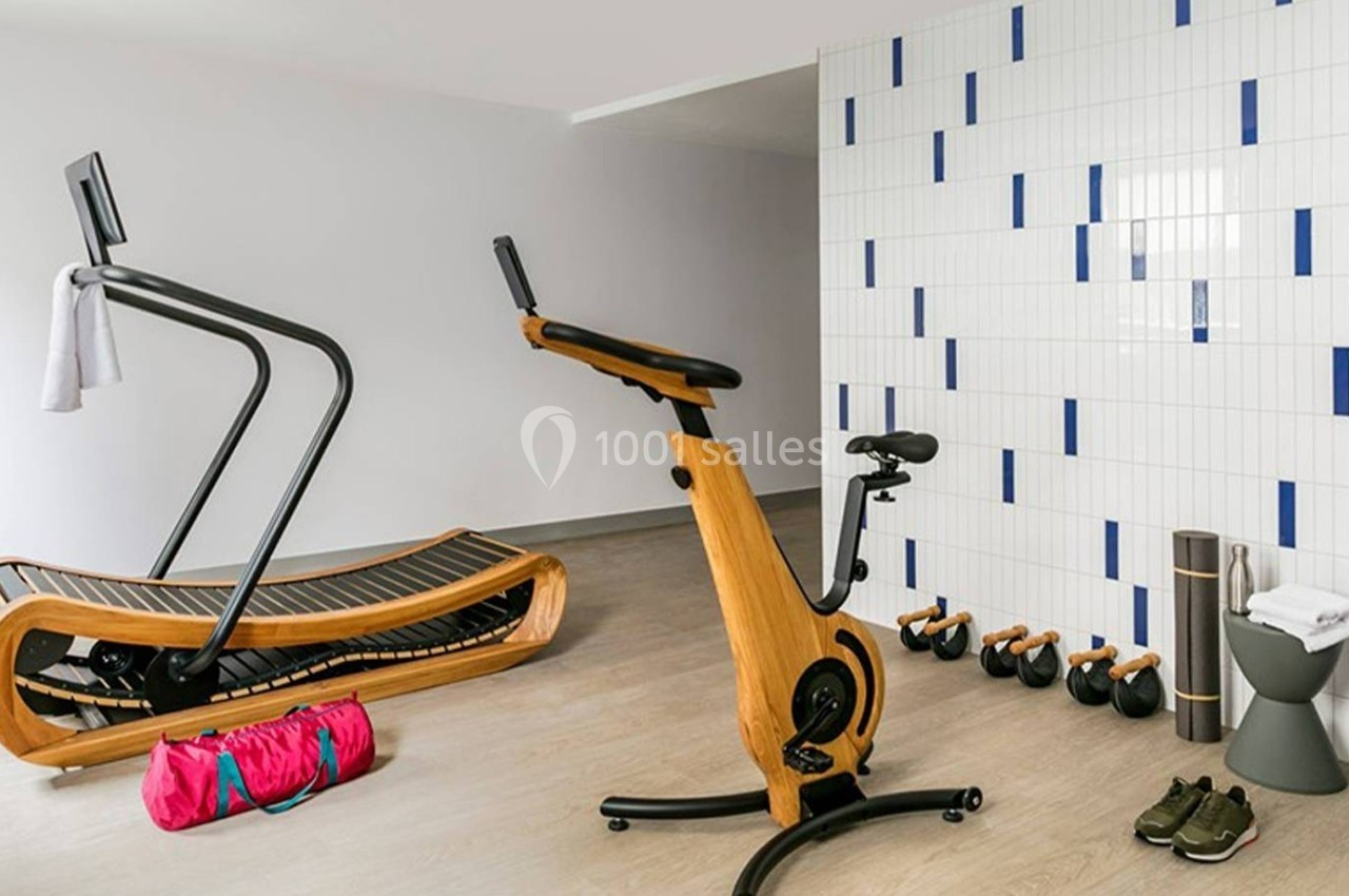 Salle de sport moderne avec tapis de course en bois, vélo d'exercice, haltères, sac de sport et chaussures au sol.