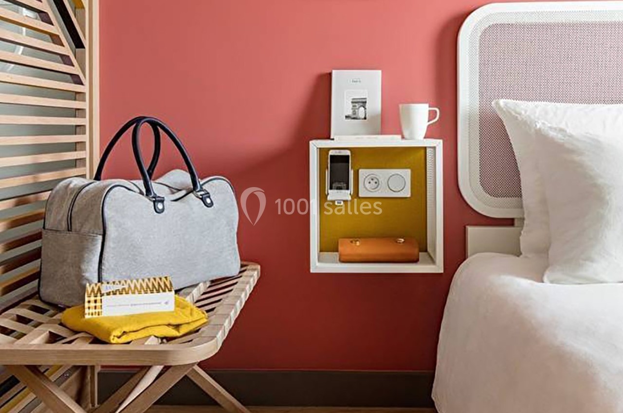 Sac de voyage gris posé sur un banc en bois près d'un lit, avec une étagère murale contenant des objets.