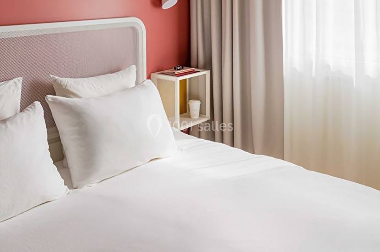 Lit avec draps et oreillers blancs, tête de lit en métal, mur rouge, rideaux beiges et lumière naturelle.