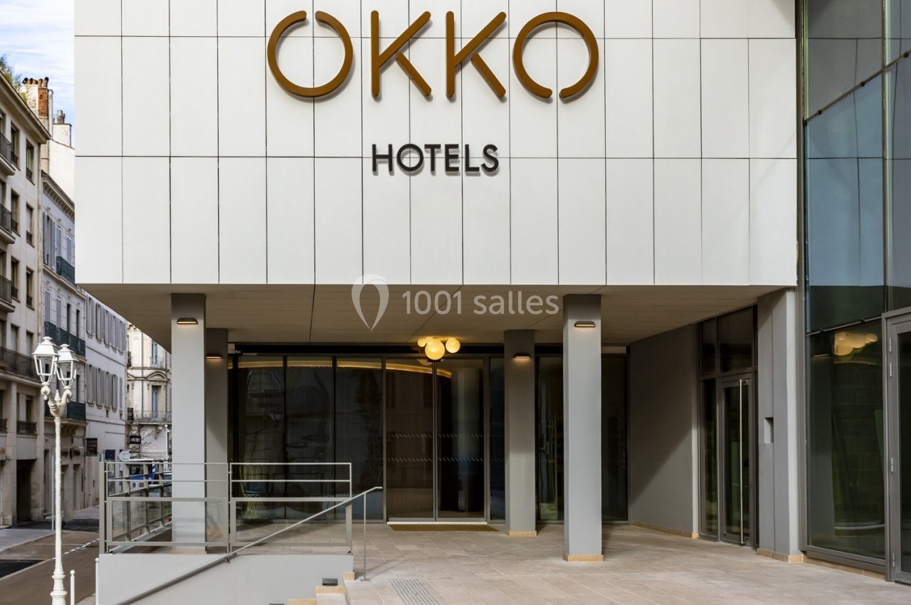Entrée moderne d'un hôtel avec une façade blanche, un logo ’OKKO HOTELS’ et une rampe d'accès.