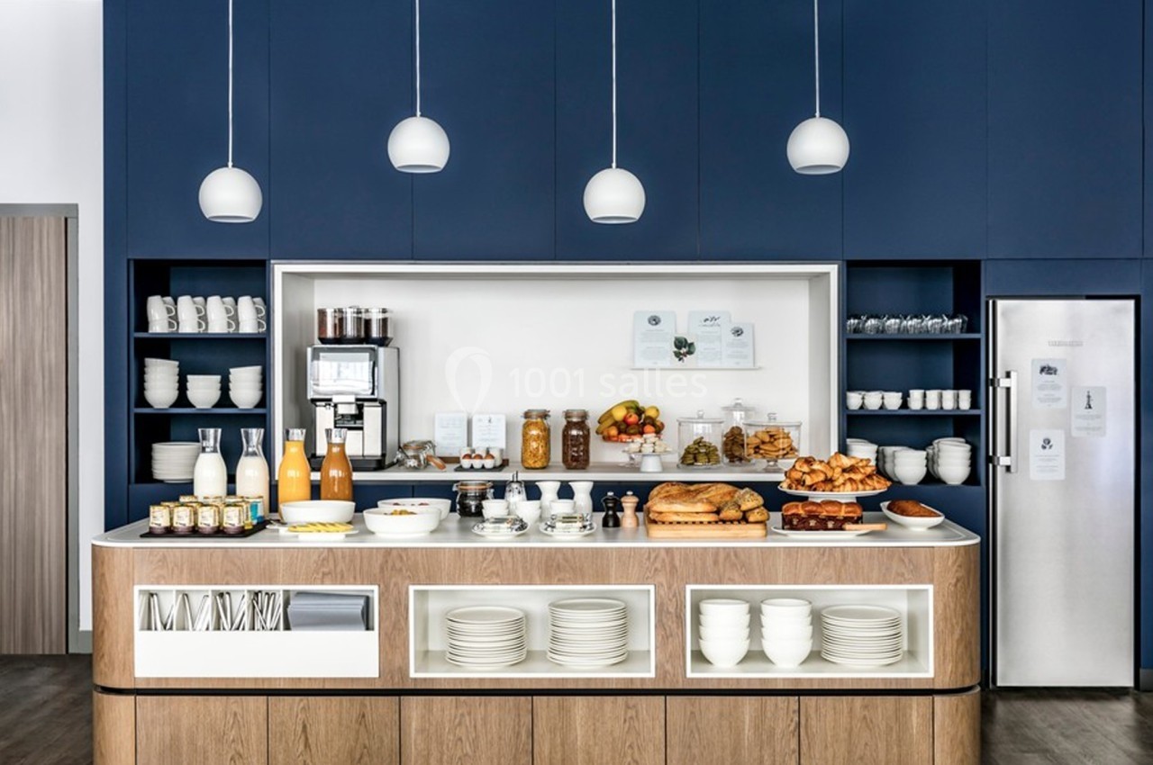 Buffet de petit-déjeuner avec boissons, viennoiseries, fruits et vaisselle, dans une cuisine moderne aux tons bleus et bois.