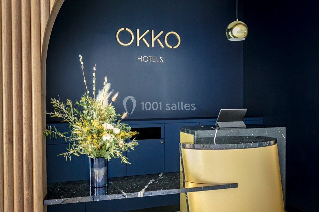 Accueil d’un hôtel avec un comptoir moderne en marbre, une décoration florale et un mur bleu portant le logo ’OKKO HOTELS’.