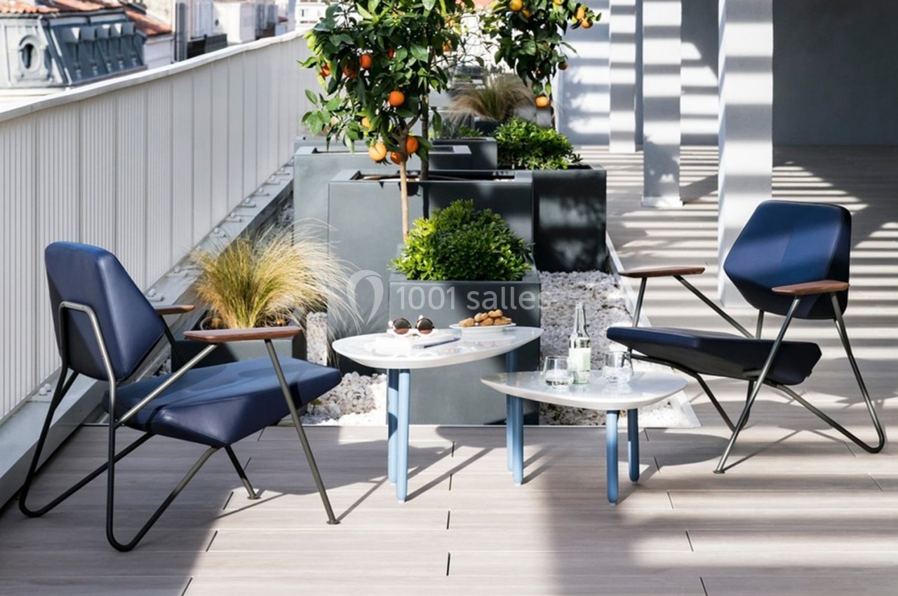 Deux fauteuils bleus entourent des tables basses blanches sur une terrasse avec plantes et oranger en pot.