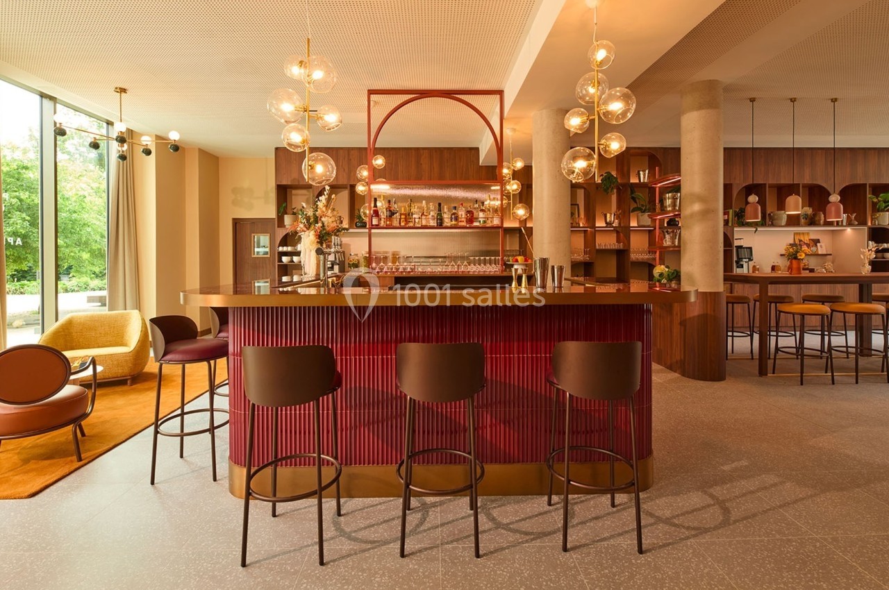 Bar moderne avec comptoir rouge, tabourets noirs, éclairage suspendu et grandes fenêtres laissant entrer la lumière…