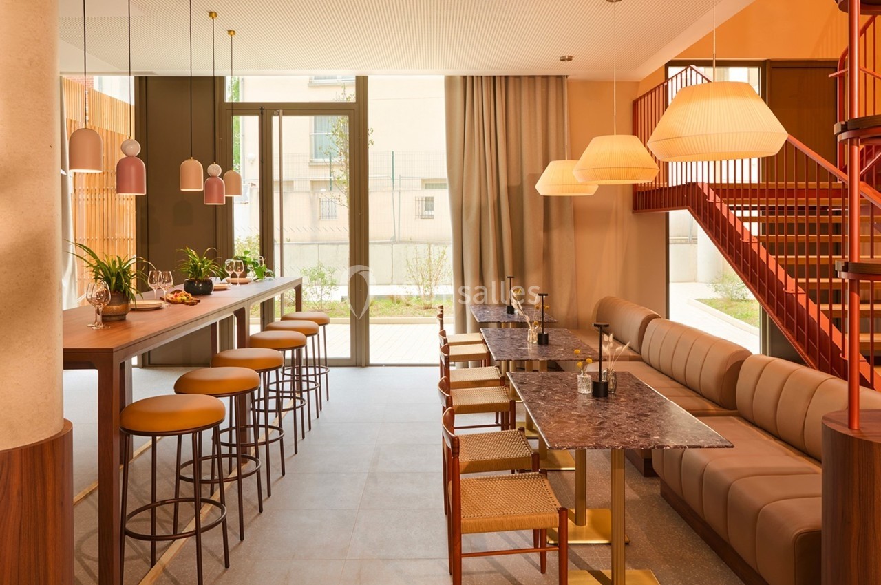 Espace intérieur lumineux avec tables, chaises, comptoir, luminaires suspendus et escalier rouge en arrière-plan.