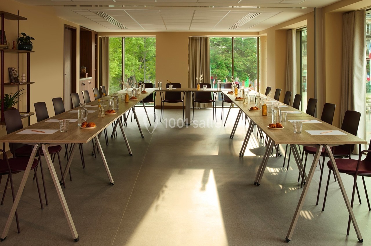 Salle de réunion lumineuse avec deux longues tables en U, chaises, bouteilles d'eau et fenêtres donnant sur un espace vert.