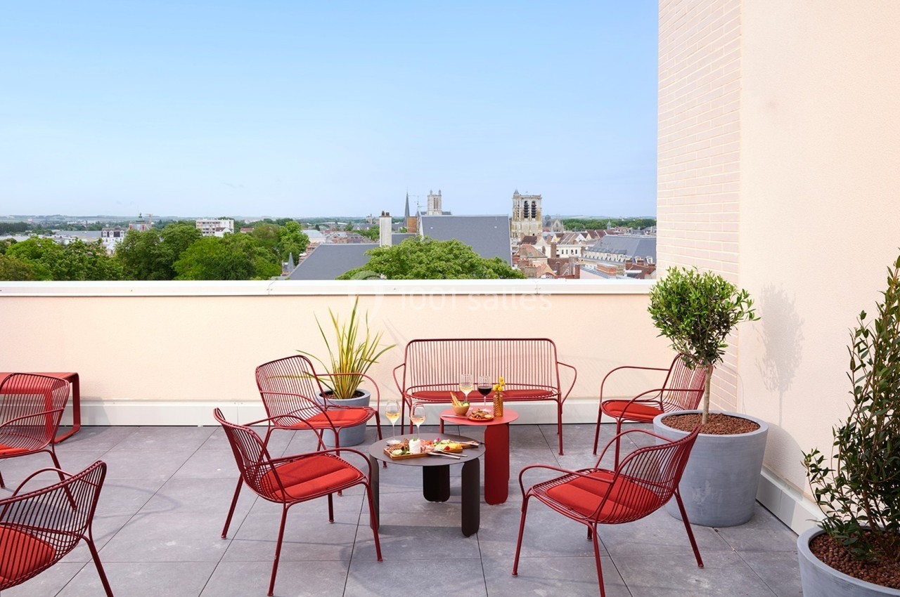 Terrasse avec mobilier rouge, table basse garnie de boissons et snacks, vue dégagée sur des bâtiments et arbres.