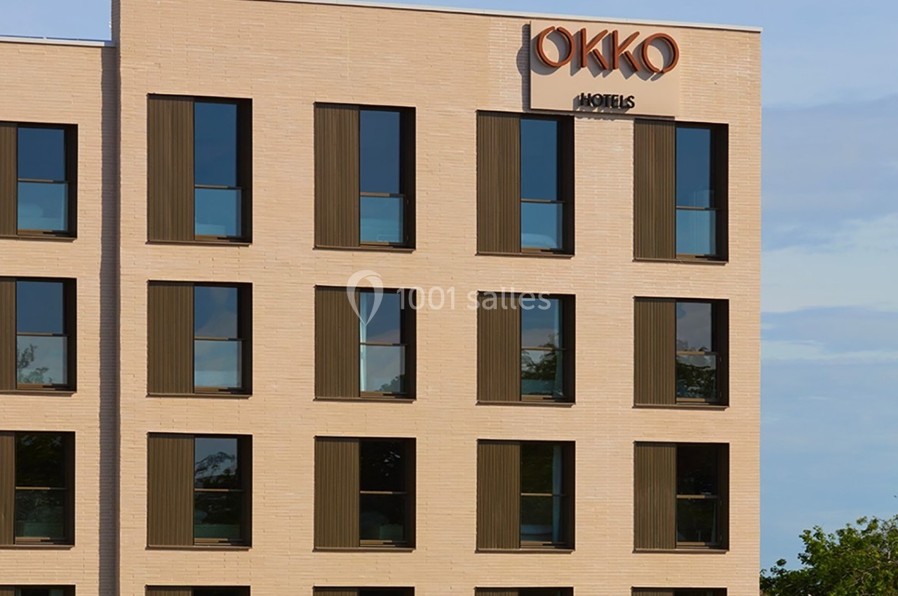 Façade d'un bâtiment moderne en briques claires avec des fenêtres rectangulaires et une enseigne ’Okko Hotels’.