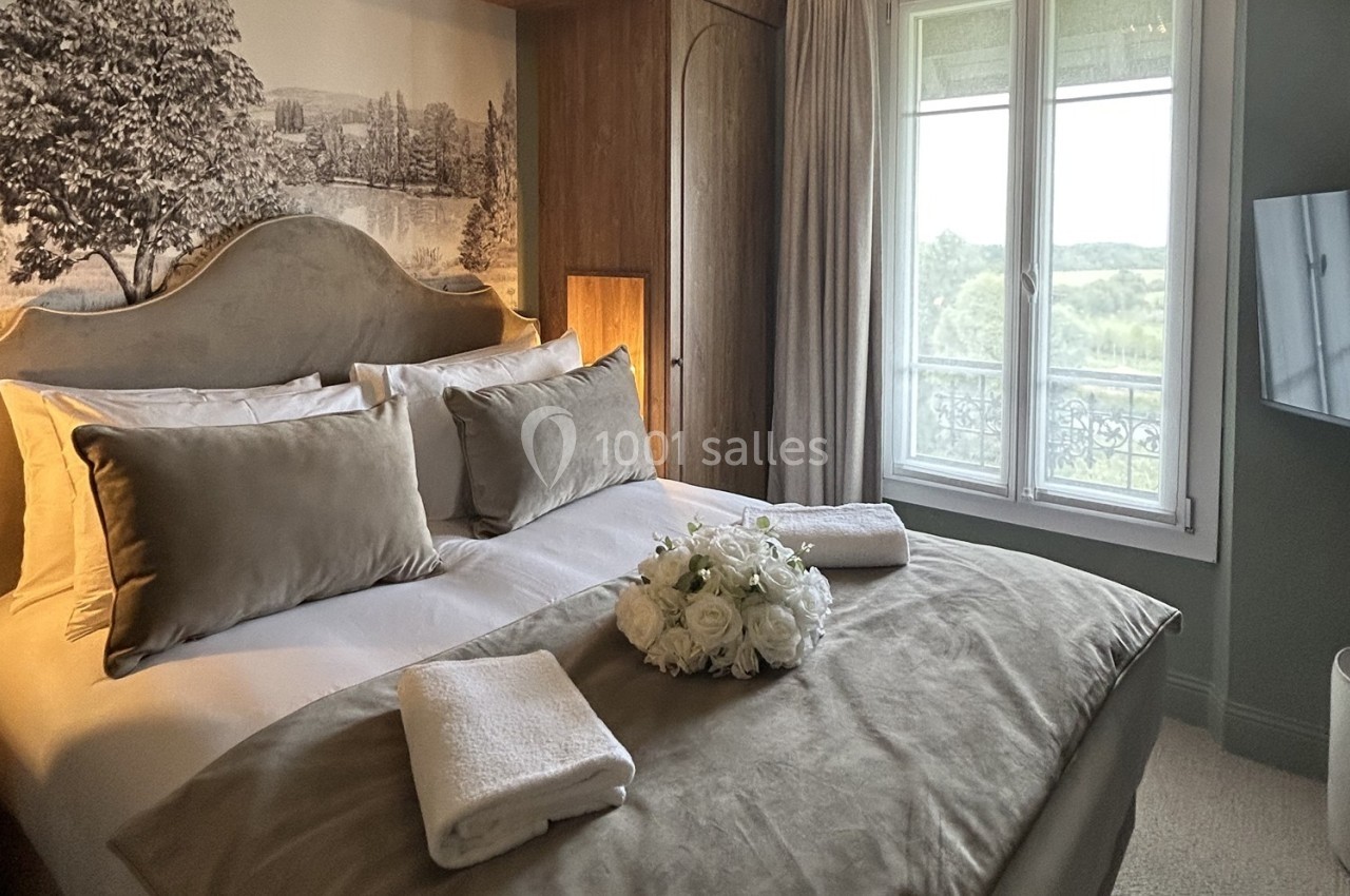 Chambre lumineuse avec lit double, coussins beiges, bouquet de fleurs blanches et vue sur un paysage verdoyant par la…