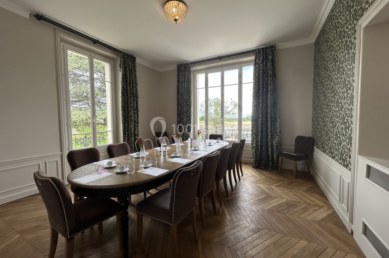 Salle à manger lumineuse avec une grande table ovale dressée, entourée de chaises, et vue sur un jardin à travers de grandes…