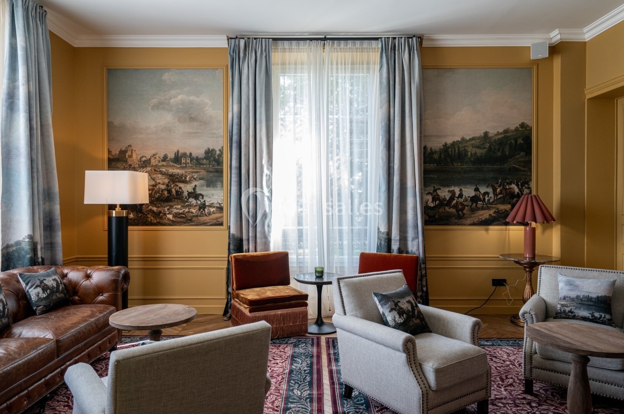 Salon élégant avec fauteuils, canapé en cuir, tables basses, rideaux et peintures murales représentant des paysages.