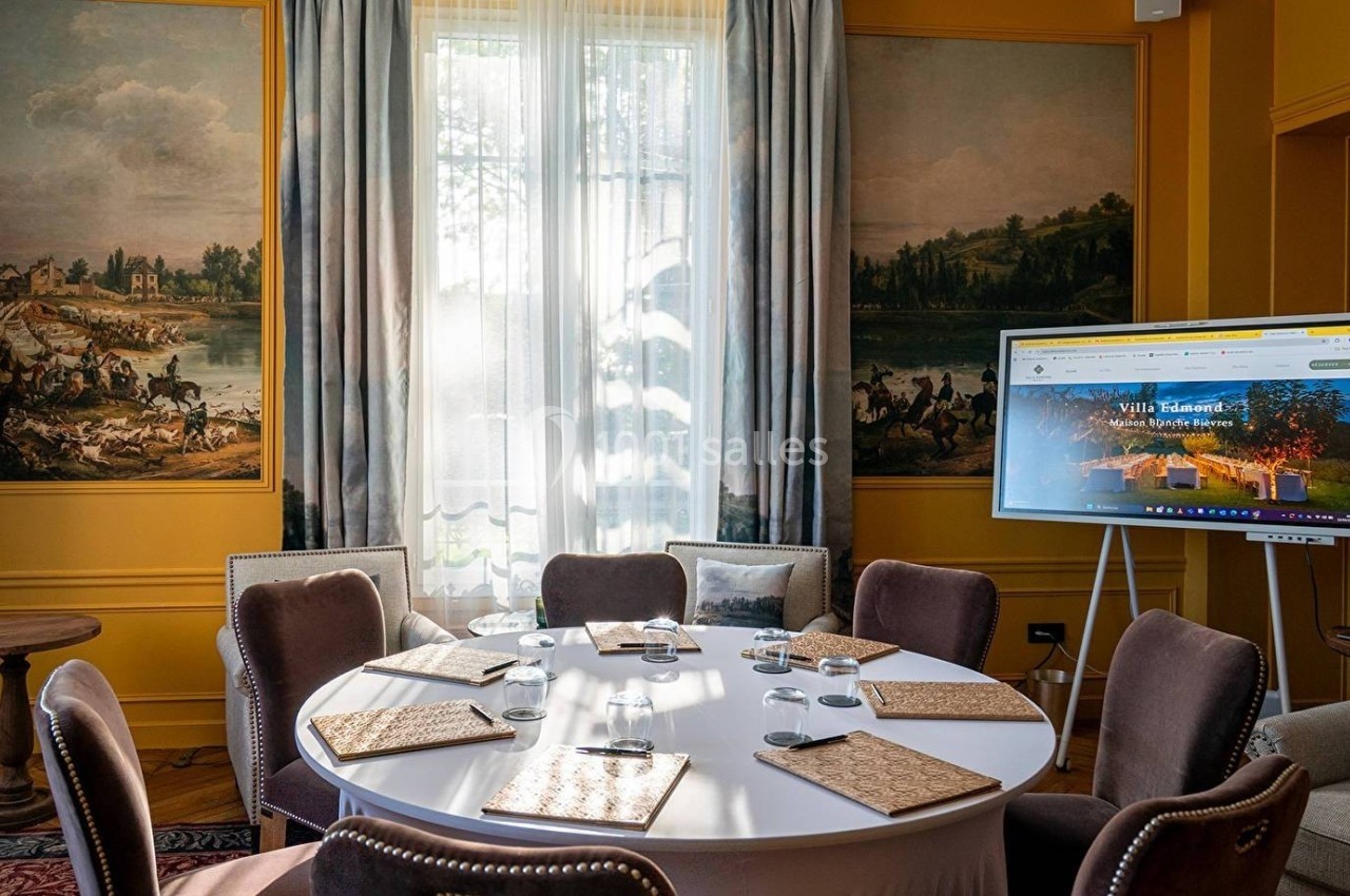 Salle de réunion élégante avec table ronde, chaises, écran de présentation et décoration murale classique.
