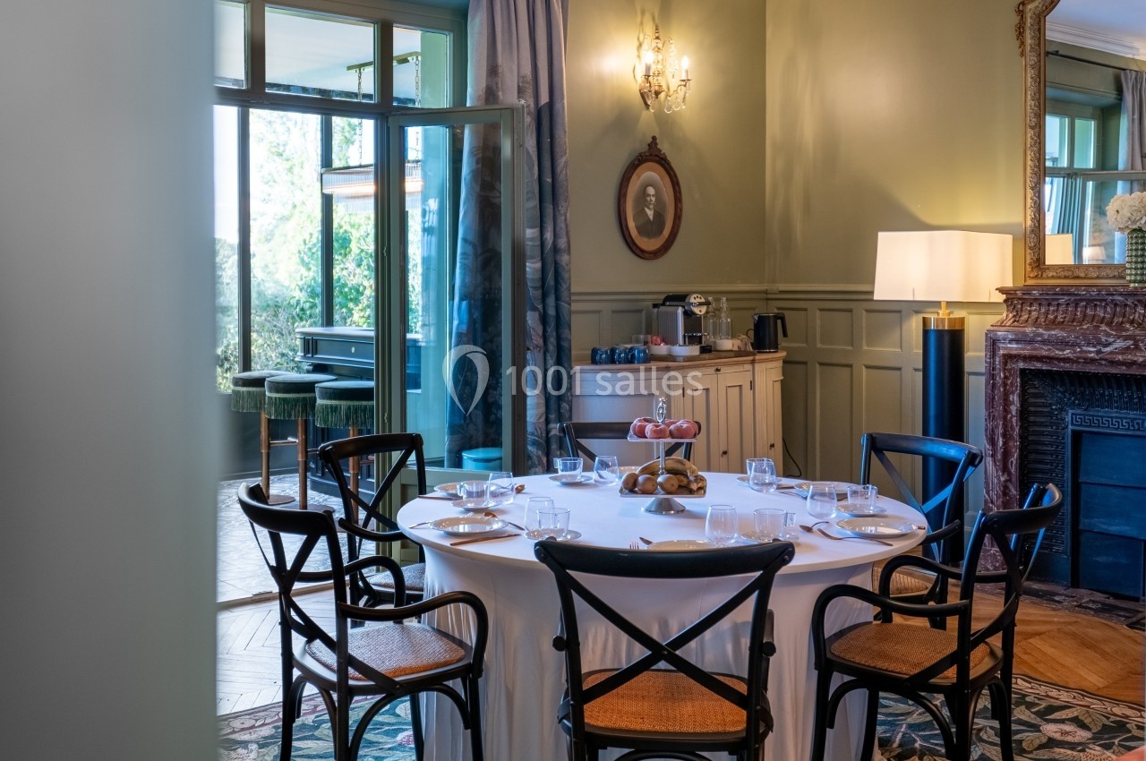 Salle à manger élégante avec table ronde dressée, chaises en bois, buffet et lumière naturelle entrant par une baie vitrée.