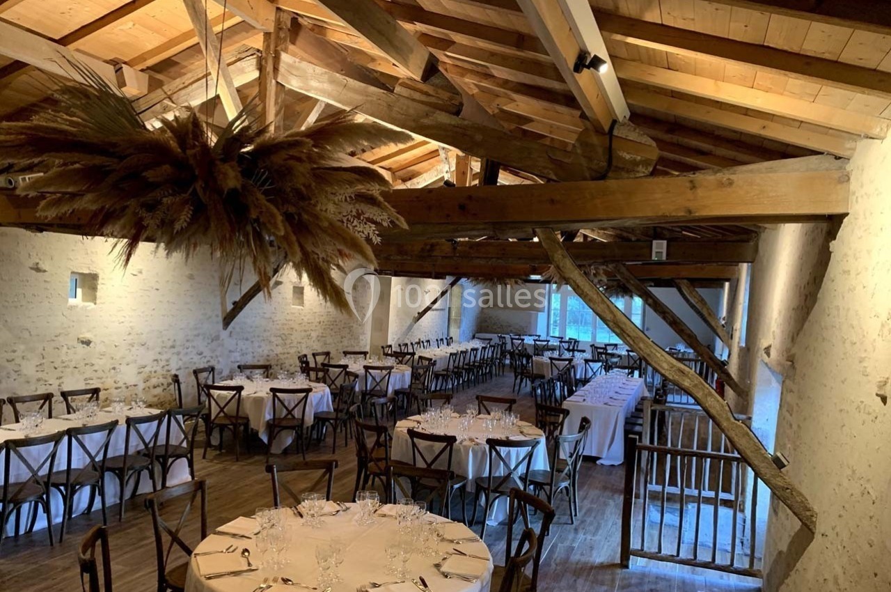 Salle de réception avec poutres apparentes, tables rondes dressées et décoration suspendue en herbes séchées.