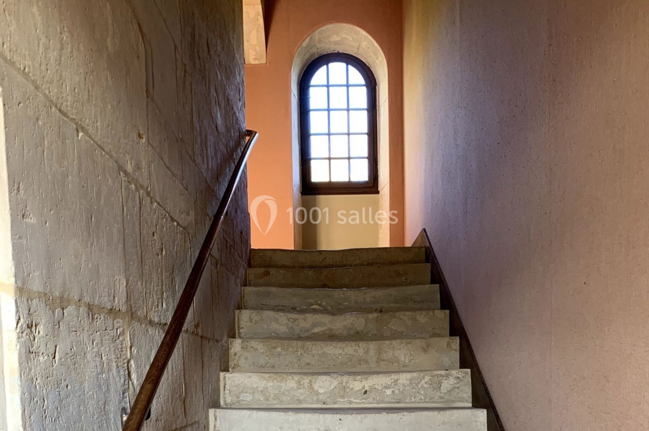 Escalier en pierre avec rampe en métal menant à une fenêtre cintrée laissant passer la lumière naturelle.