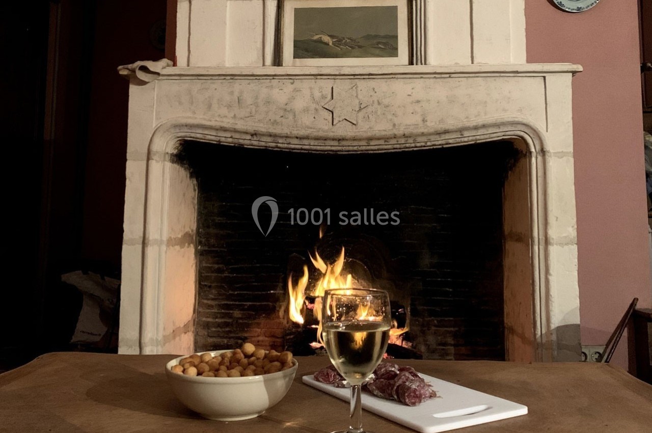 Verre de vin, bol de cacahuètes et charcuterie sur une table devant une cheminée allumée dans un intérieur chaleureux.
