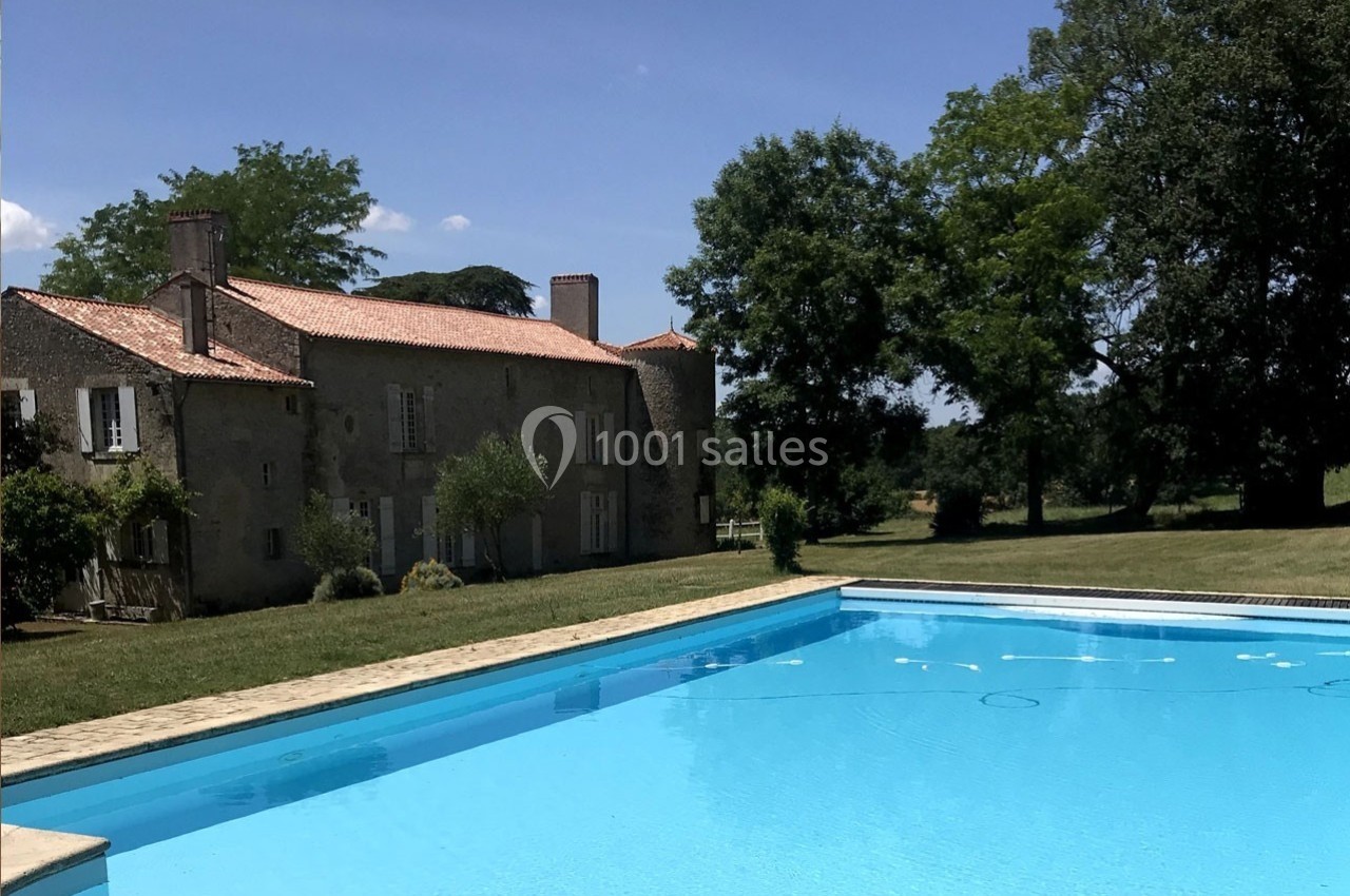 Grande piscine extérieure devant une maison en pierre avec toit en tuiles, entourée d'arbres et de pelouse.