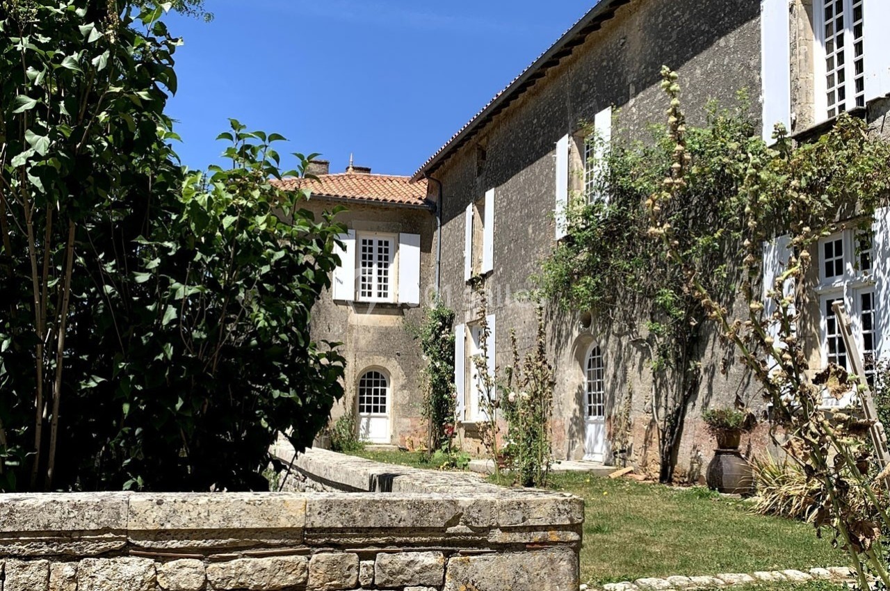 Façade d'une maison en pierre avec volets blancs, entourée de végétation sous un ciel bleu.